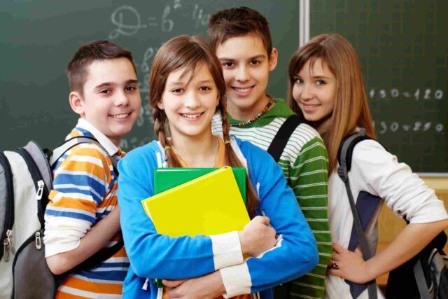 A group of four students with backpacks and books standing together in front of a chalkboard.