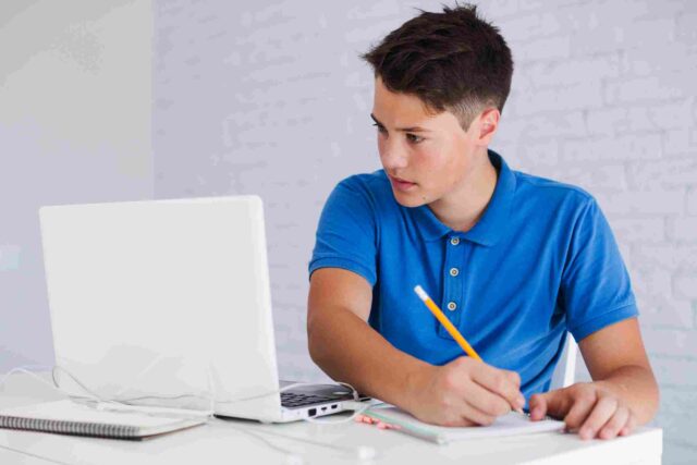 A focused teenage boy using a laptop and notebook for virtual study and effective online revision in preparation for GCSE Exams in Year 11.
